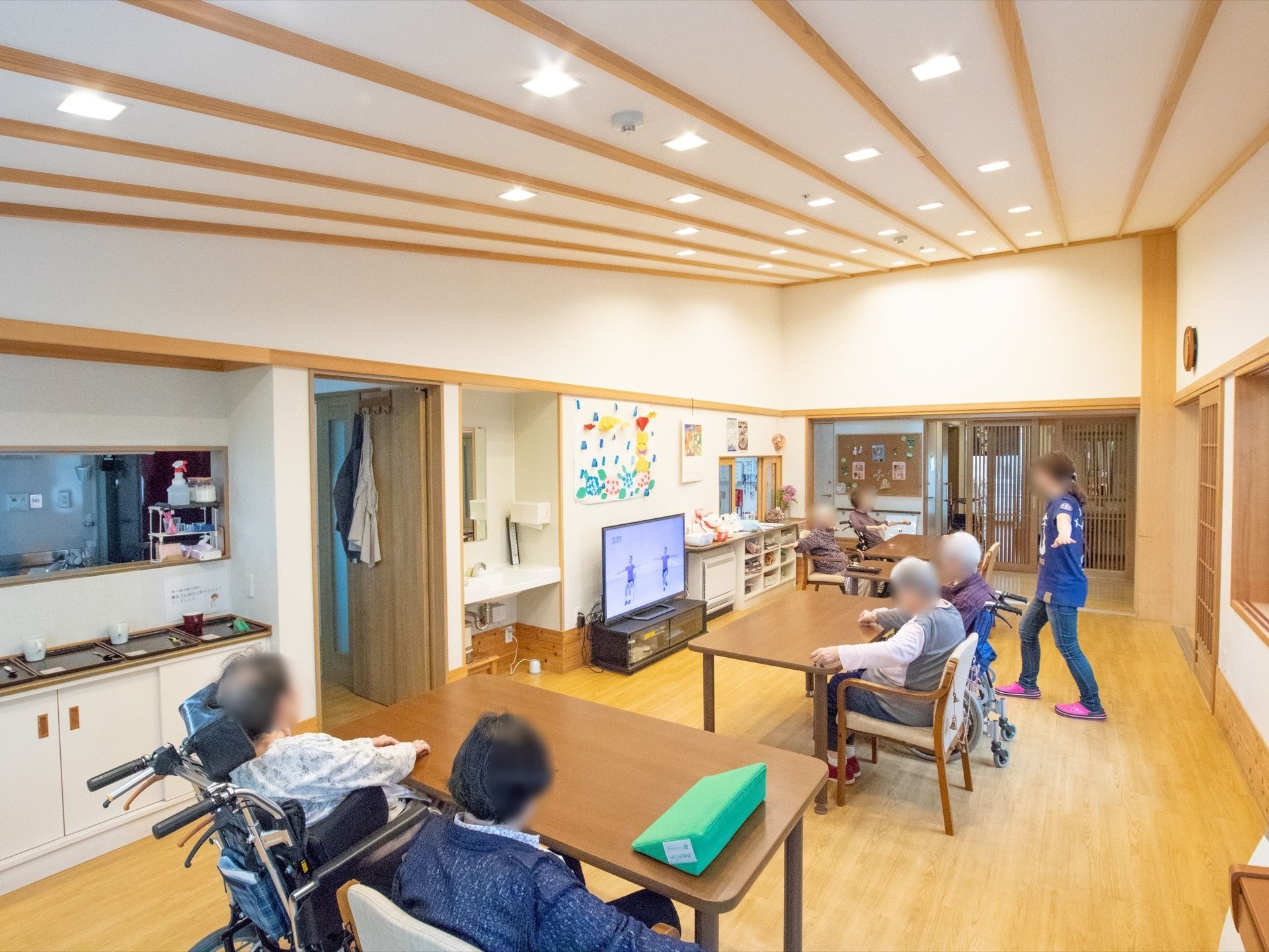 ダイニングテーブルに入居者様が2人ずつ腰かけている。女性スタッフとテレビ画面で映像を見ながら手を伸ばす運動を行っている。