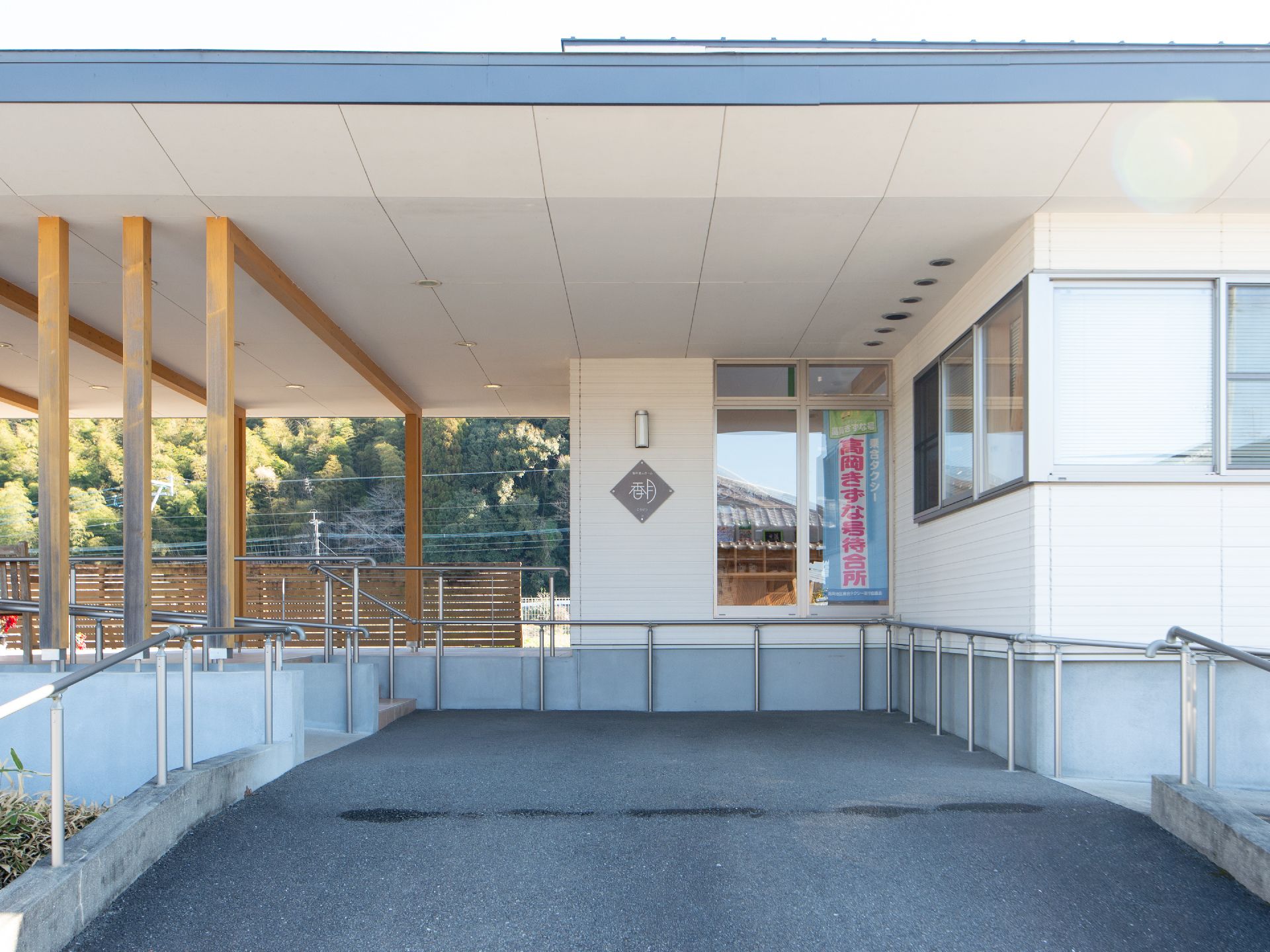 幅の広いアスファルトの斜面の先に、施設名称表示が施された壁面が見える。建物の奧には木々の緑が広がっている。