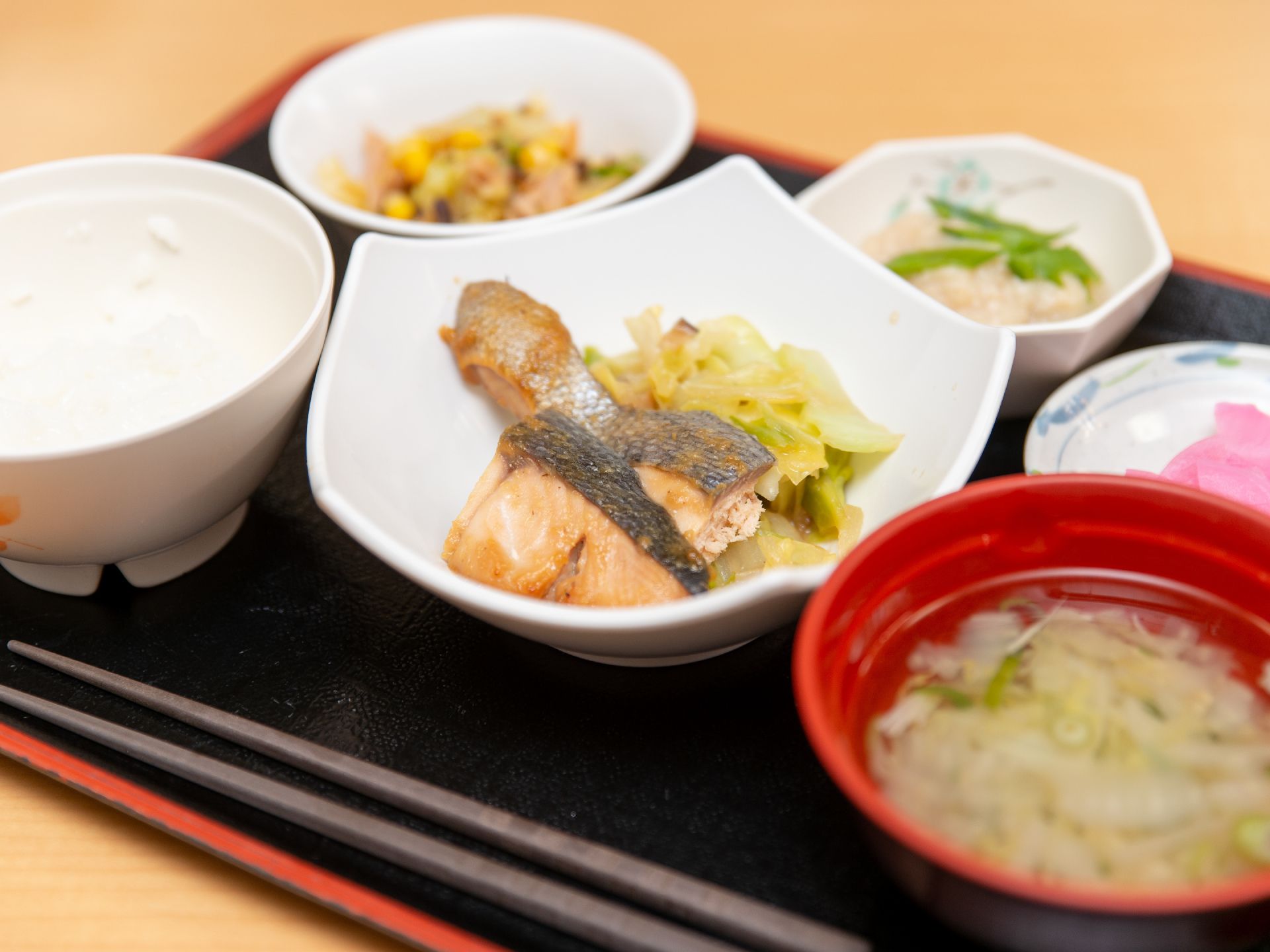 黒色のプレートに、ご飯、みそ汁の他、おかずが３皿と漬物がのっている。焼き魚が主菜である。手前に箸が置かれている。