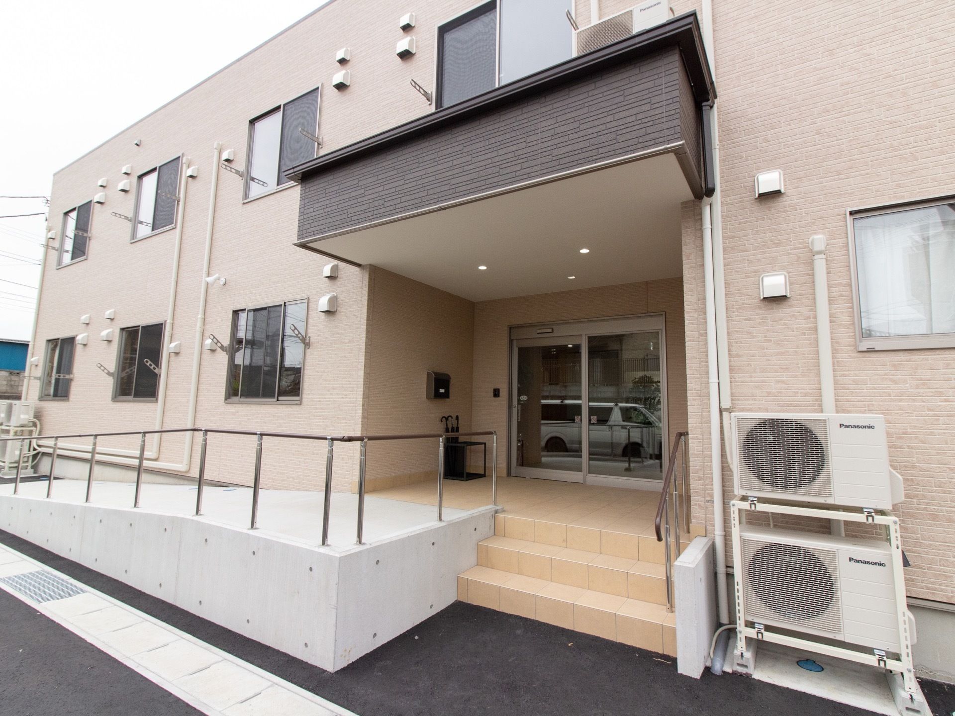 施設はブラウン色の外壁の建物になっており、玄関まではスロープと階段が設置されている。階段の脇にはエアコンの室外機がある。