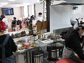 入居者様が食事をしている風景が見える位置に厨房がある。三角巾とエプロン、マスクを着用したスタッフが食事を作っている。