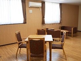 木目調の床に４人掛けテーブルと椅子、部屋の隅にはソファとテーブルが設置されている。窓が２つあり日当たりがよい。