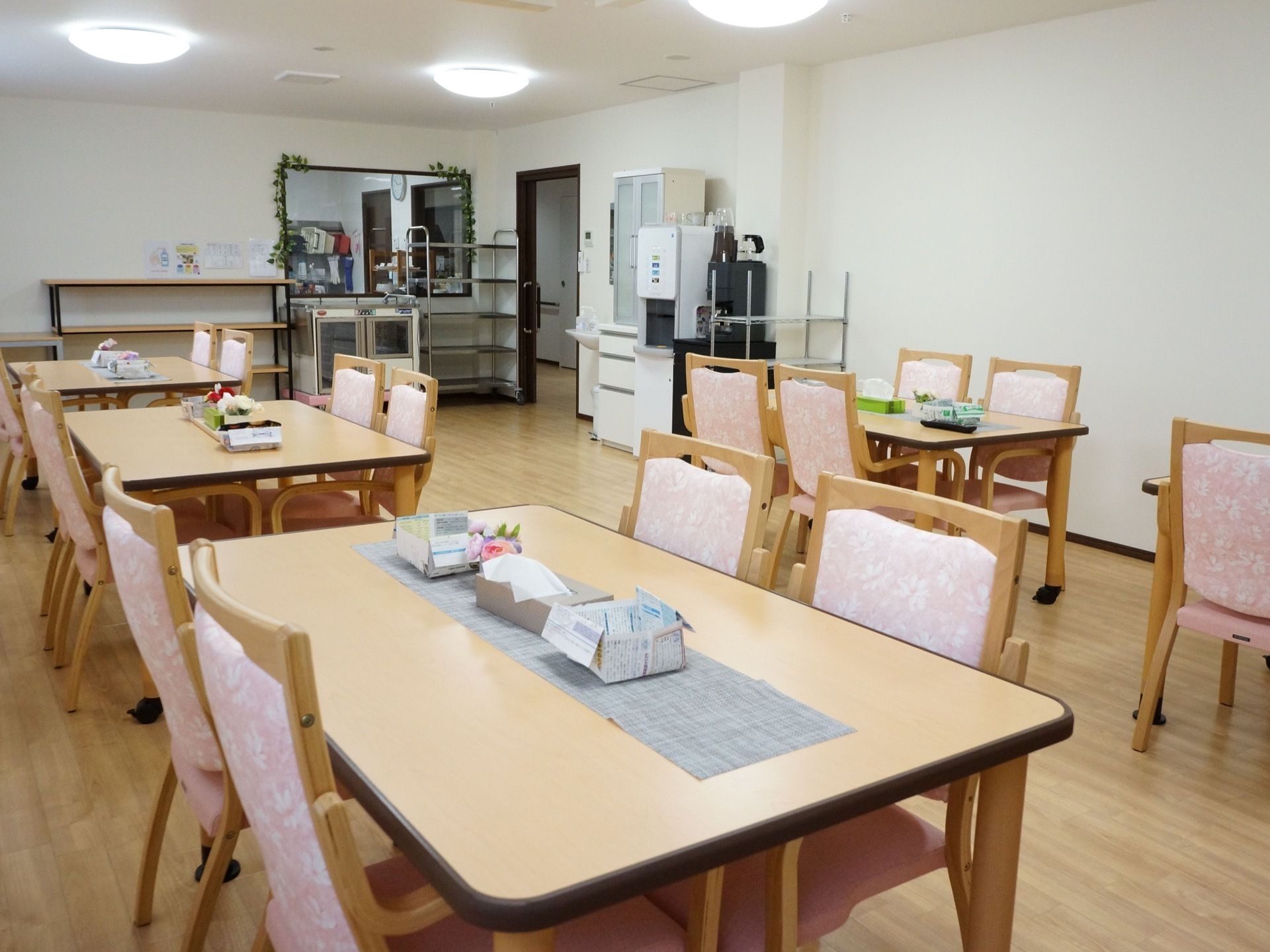 四人掛けのテーブルの上には造花やティッシュペーパーが置かれている。奥には、食事を運ぶ時に使うワゴンやウォーターサーバーがある。