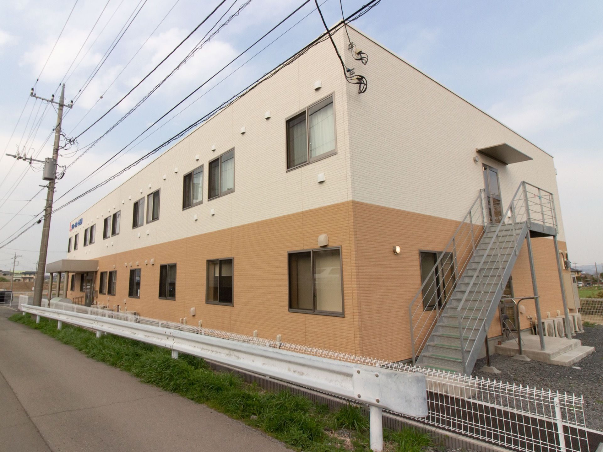 １階部分はオレンジ、２階部分は白に塗られた外観の２階建ての建物。右手前にはグレーの鉄製の外階段がある。