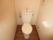 白い壁に囲まれたシンプルな洋式トイレである。タオル掛けハンガーやペーパーホルダ―が壁に設置されている。