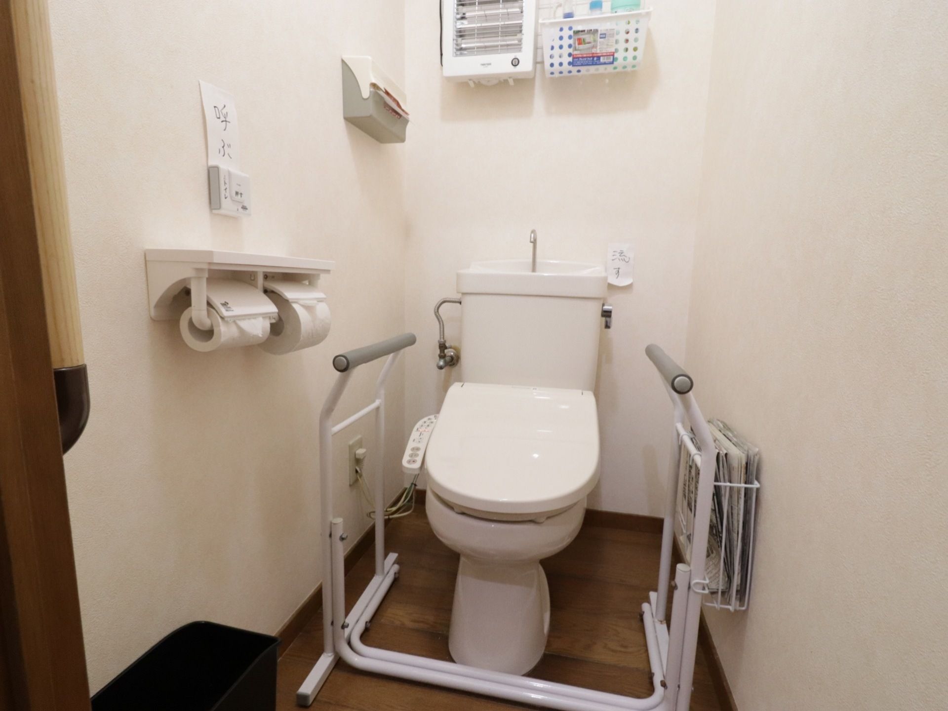 便座の左右に手すりが取り付けられている。洗浄機付きタイプのトイレで、壁には呼び出しボタンがついている。