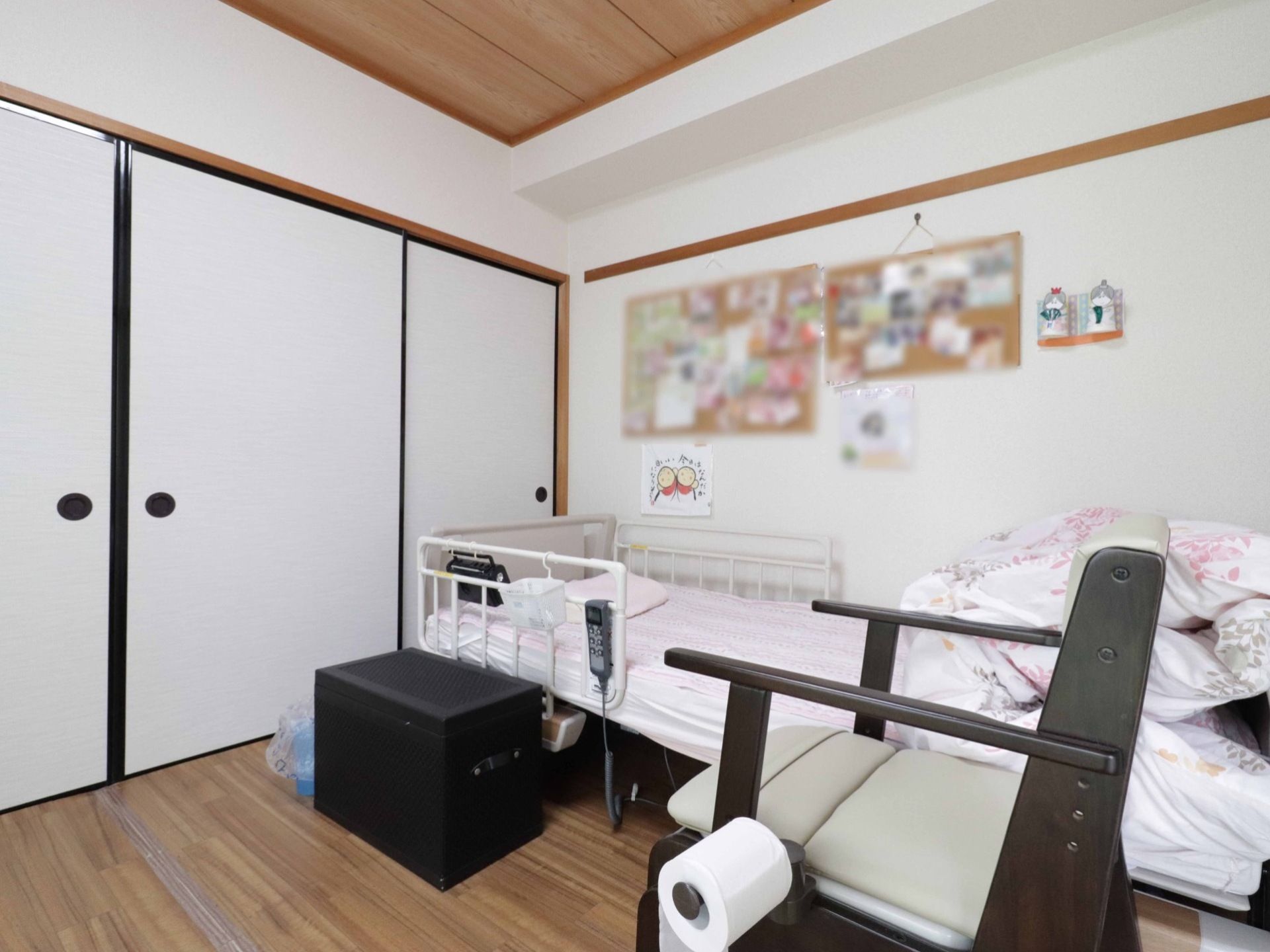 介護用のベッドと椅子、収納付きのスツールが置かれている。壁のコルクボードには入居者様の記念写真などが貼られている。