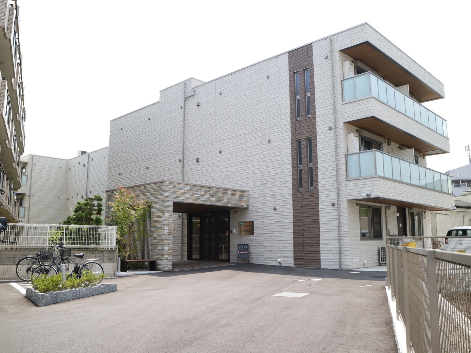 道路から奥まった場所に立地していて、周囲の通路は広々としたスペースが設けられている。白とベージュのツートンカラーが印象的な建物外観。