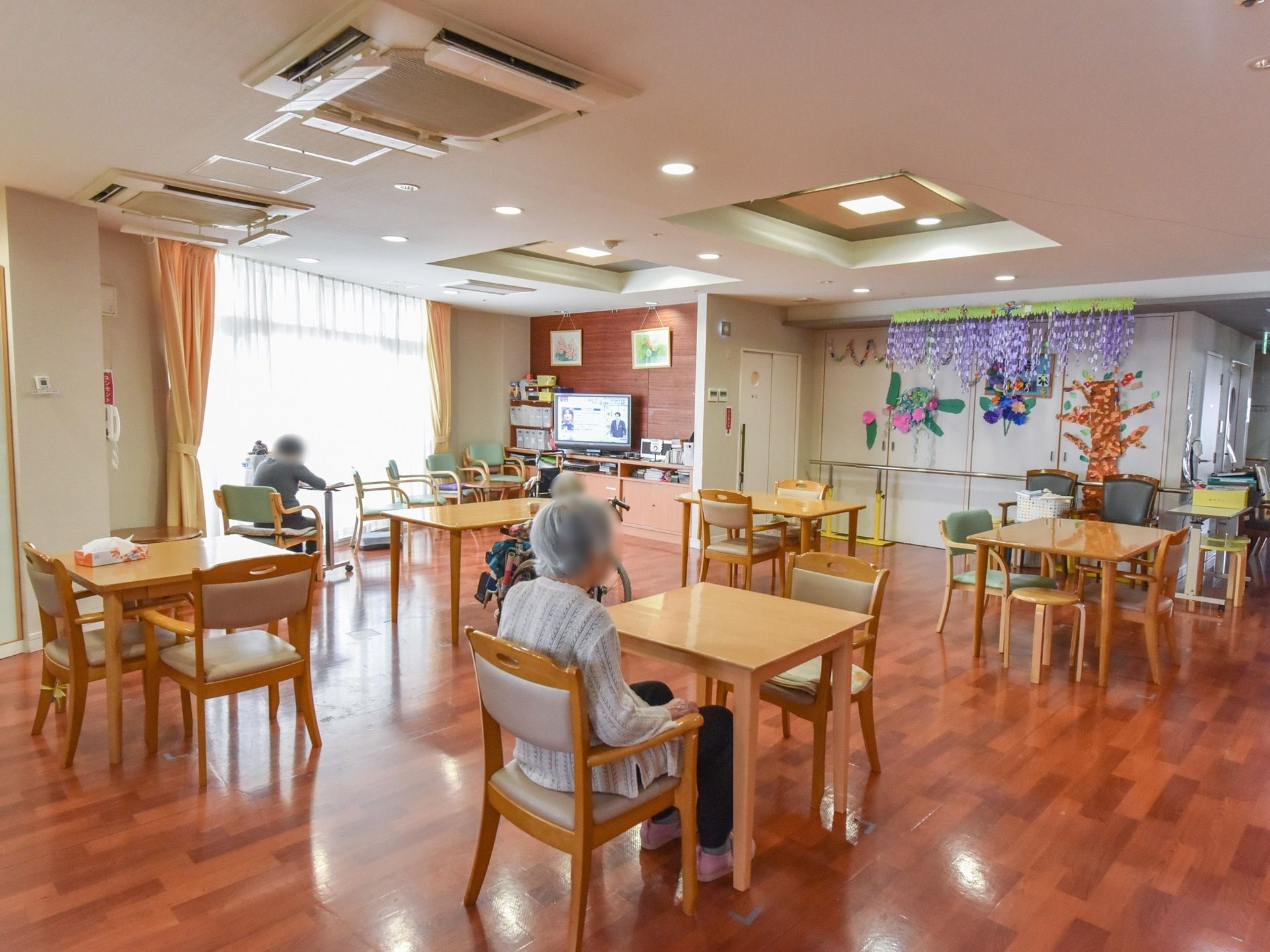 食堂はフローリングの床に、ナチュラルな色合いの木製のテーブルと椅子が並べられている。入居者様が椅子や車椅子に座っている。