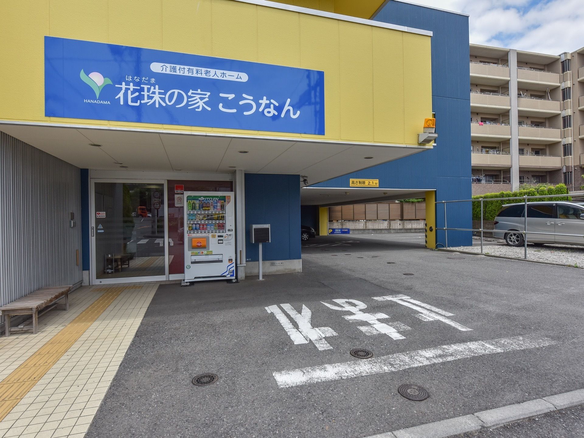 入口の上の黄色い外壁に施設名が書かれた青い看板が設置されている。入口の自動ドアの前には自動販売機が設置されている。