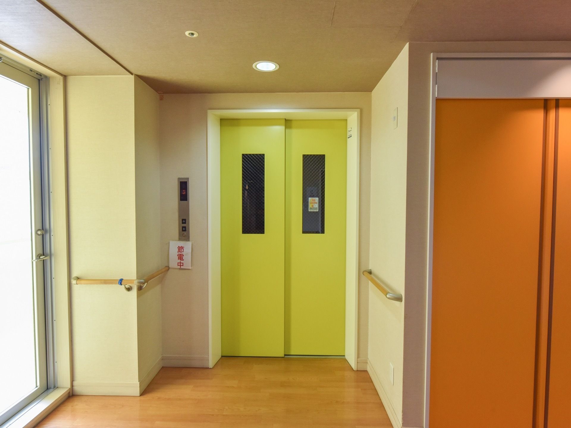 明るい黄色のドアのエレベーターが設置されている。操作ボタンの下には節電中と書かれた紙が貼られている。