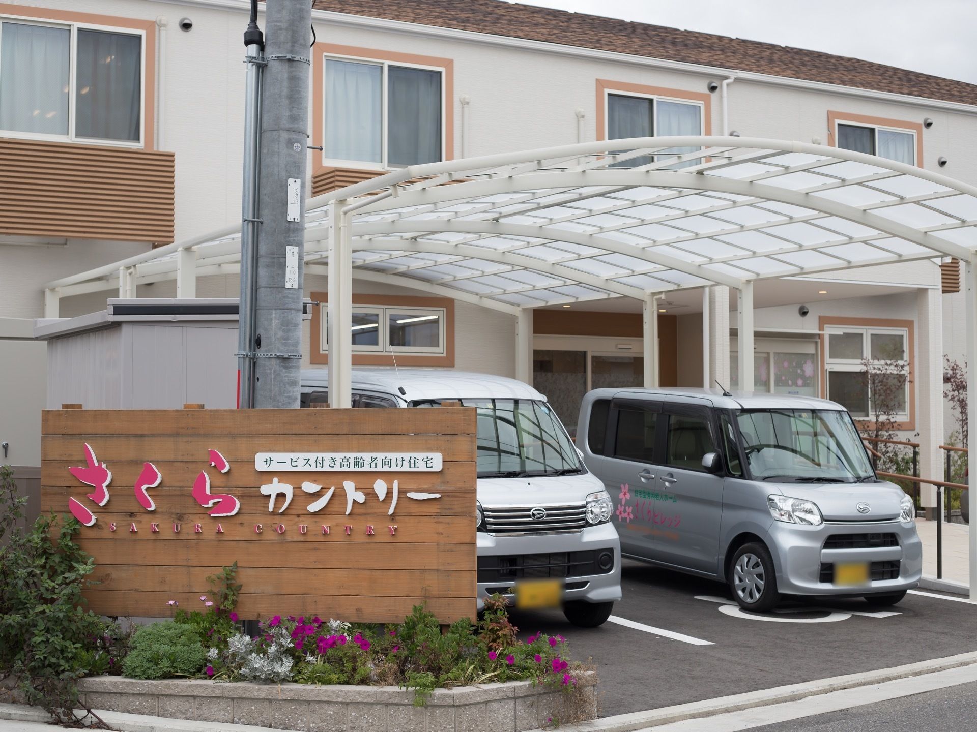 施設は2階建てのハイツのような外観である。施設の前を通る道路に面して花壇が設けられ、緑色や紫色の花々が植えられている。