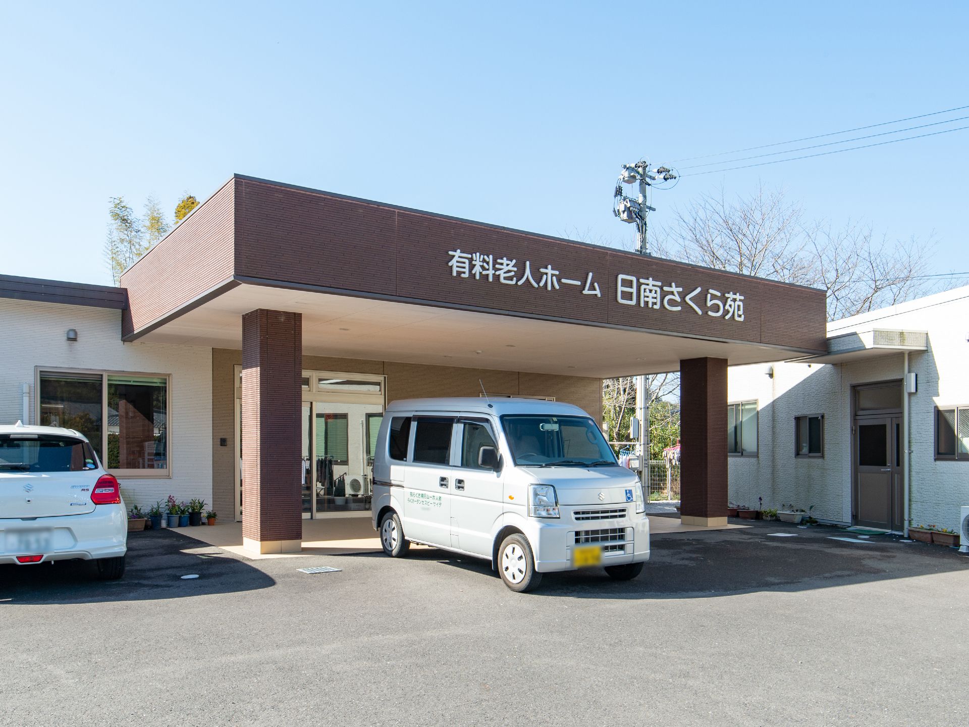 お天気の良い日の施設外観である。茶系の屋根の前面に、白い文字プレートで施設種別と名称が掲げられている。