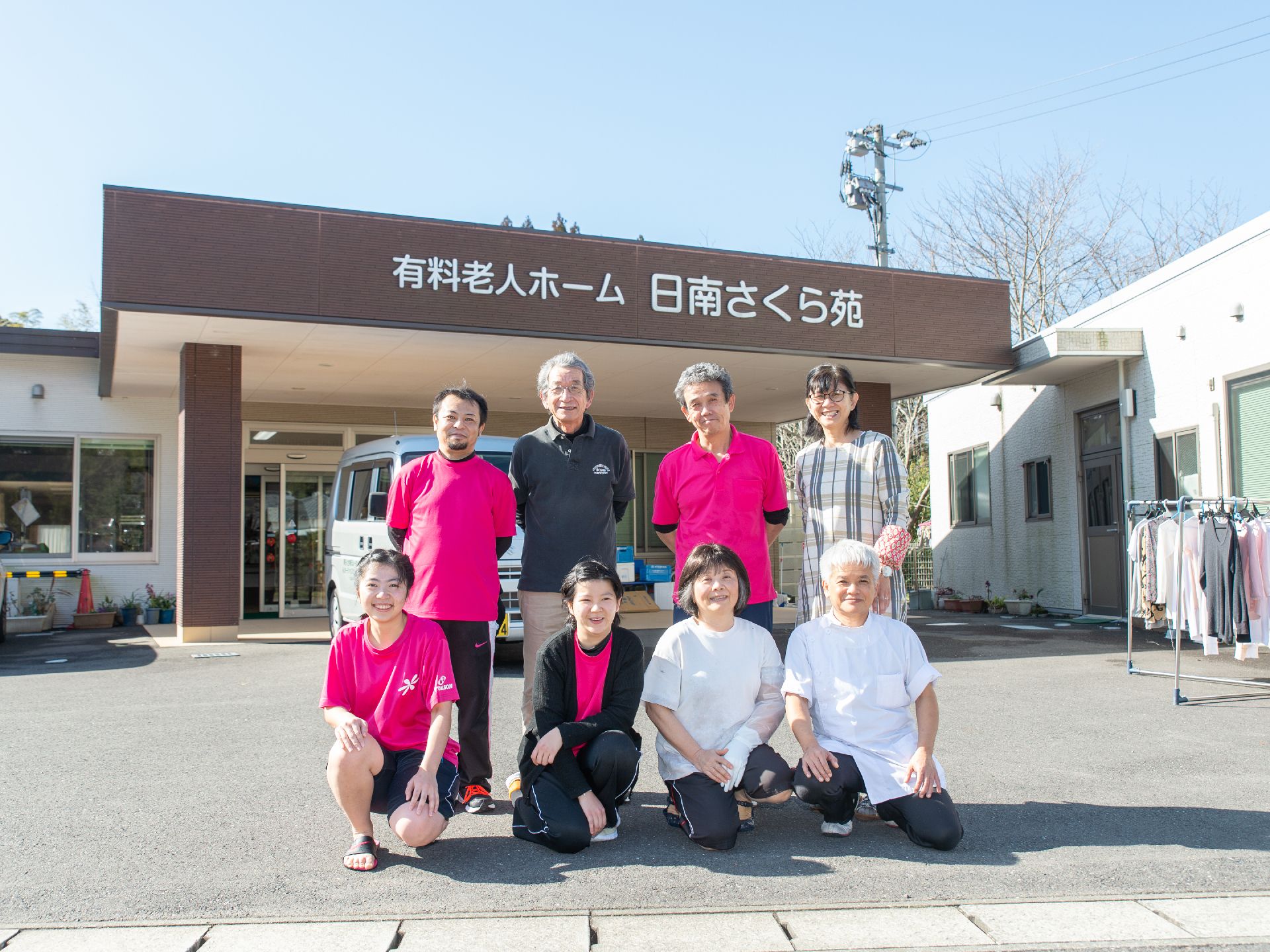 施設名が書かれた正面玄関を背に、並んで写真に納まるスタッフ