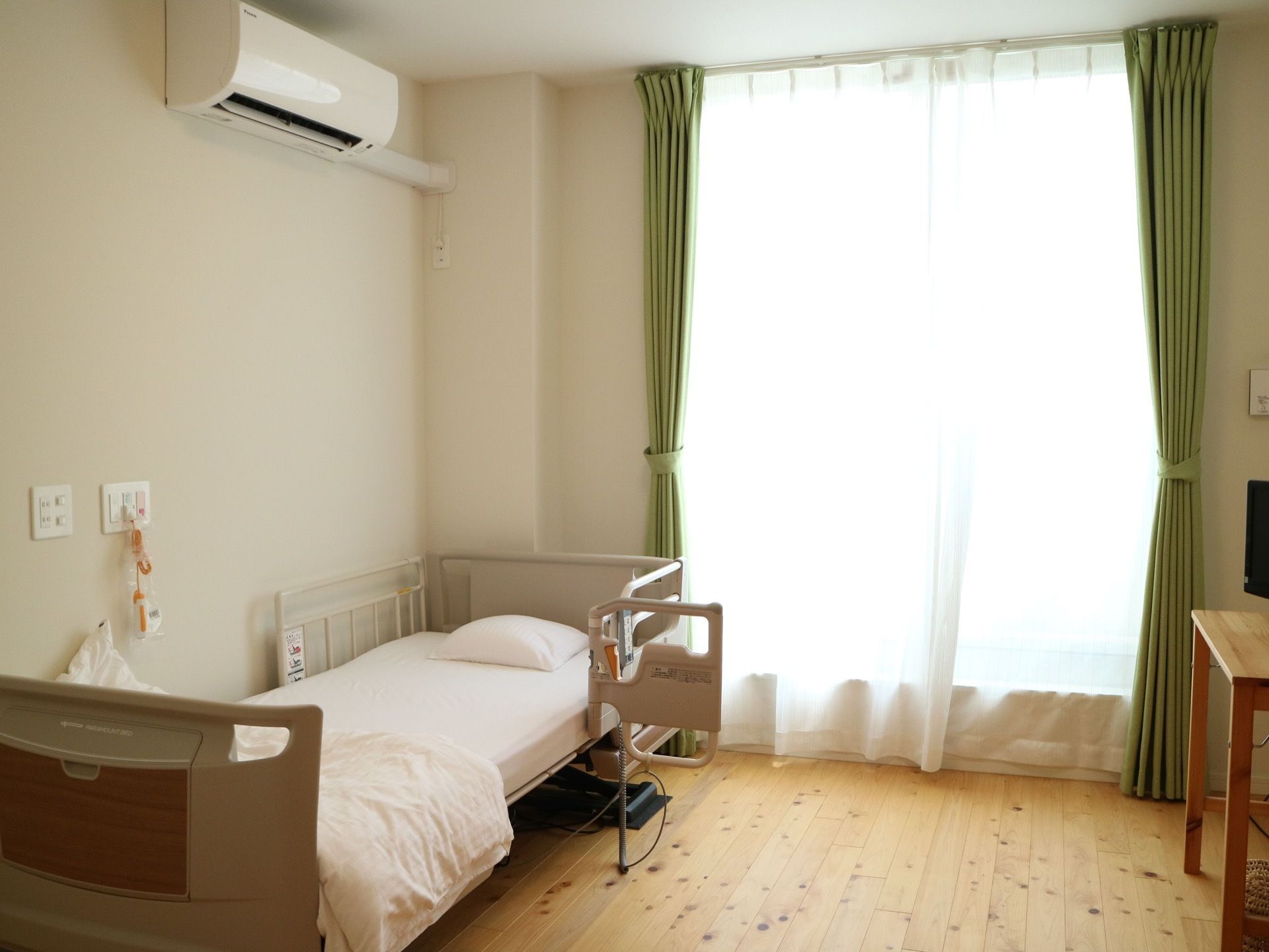 木目調の床白い壁の部屋には地窓から明るい光が差し込んでいる。エアコンや介護ベッド、テレビなどがある。
