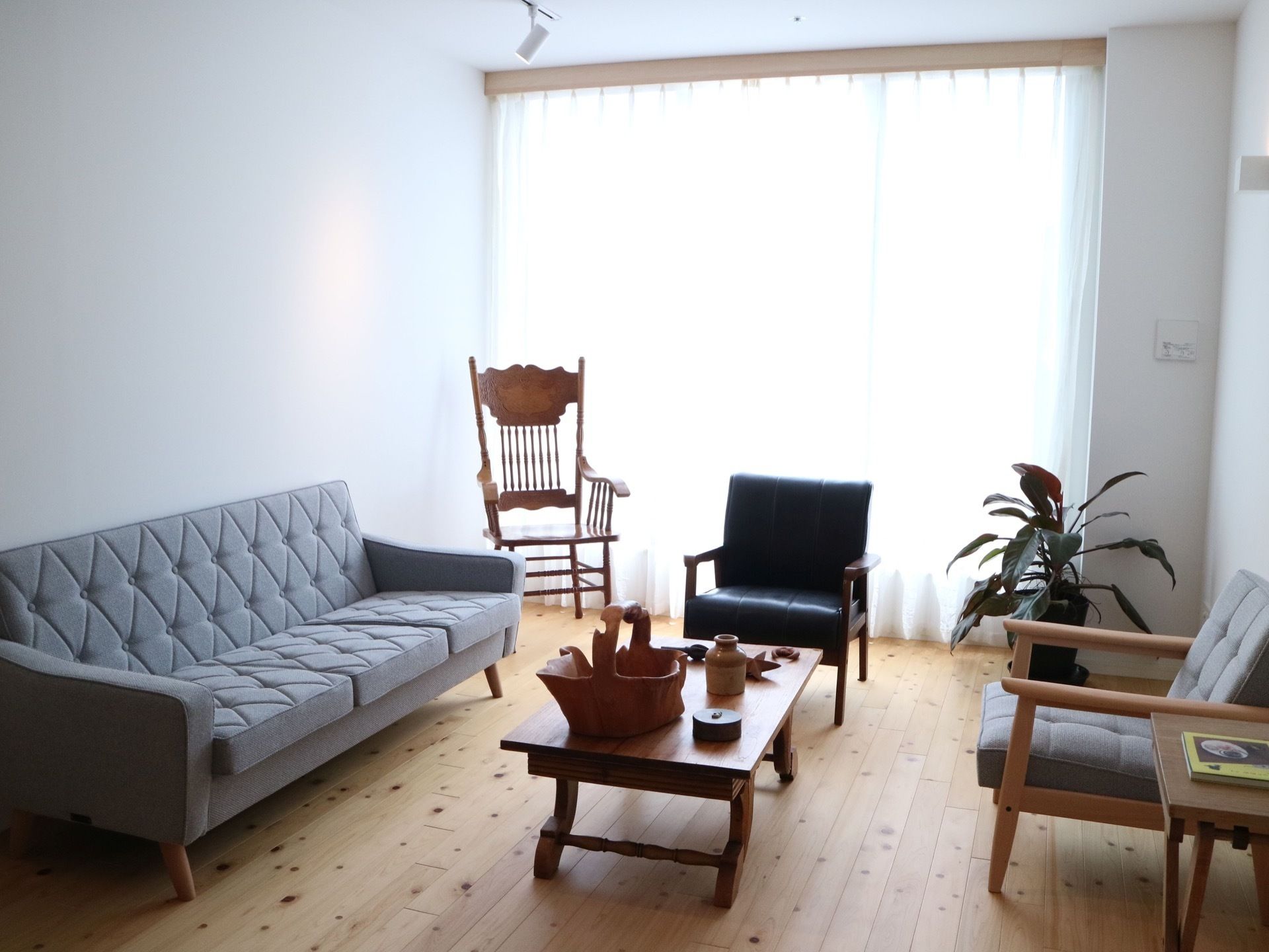 大きな地窓のある木目の床の部屋に、ゆったりと応接セットが置かれている。窓際には観葉植物と木製の椅子がある。