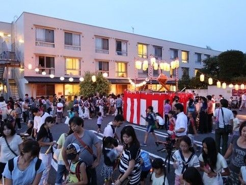 施設の駐車場で祭りが催されている。中心に太鼓が設置され、取り囲むように近所の子供や大人達も多く祭りに参加している。