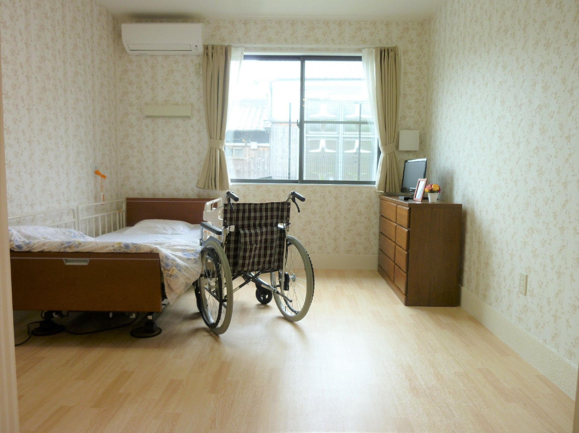 左側には介護用ベッドがあり、その右隣には車椅子が置かれている。中央奥には窓があり、その左上側にはエアコンが付いている。