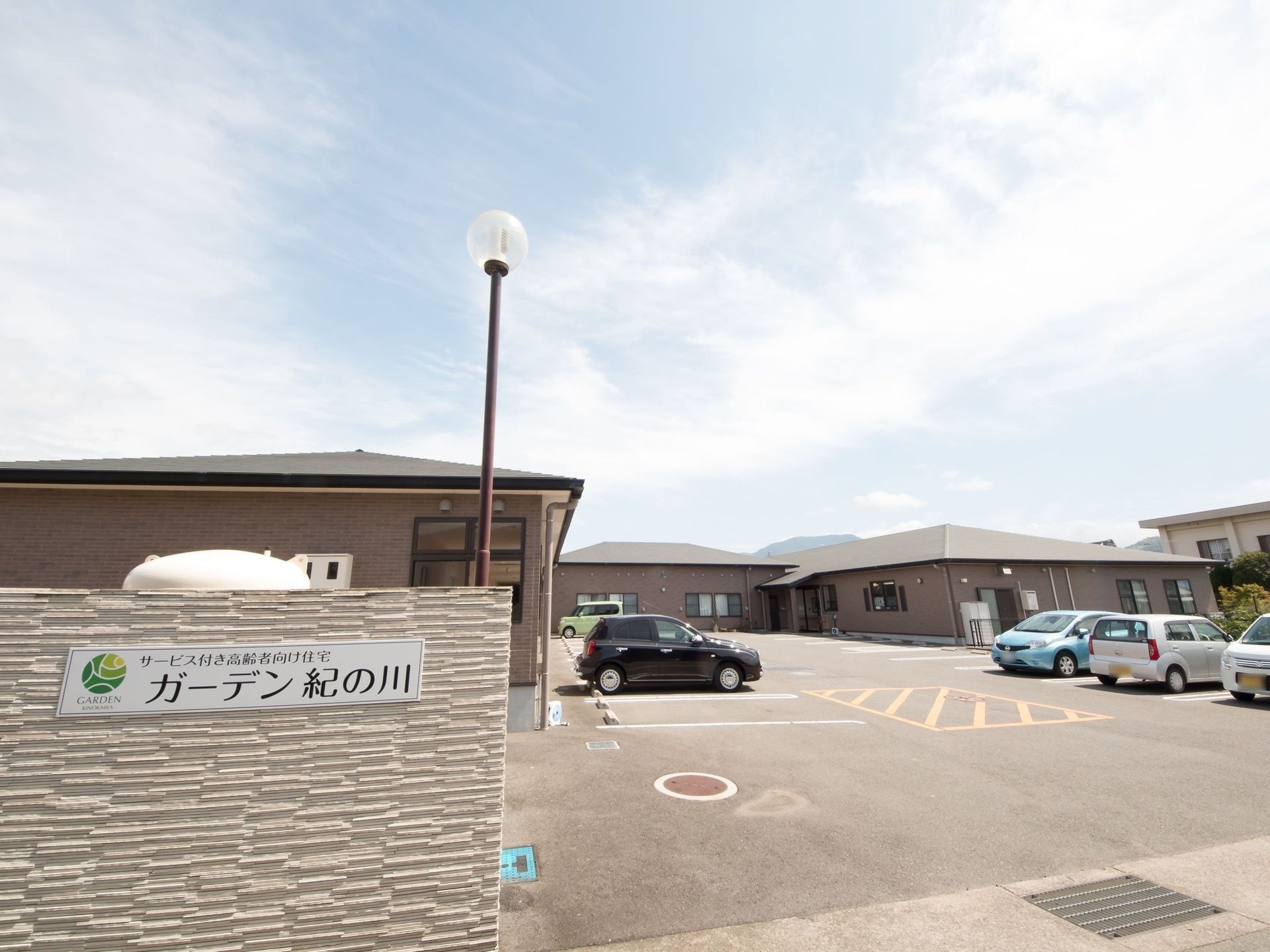手前の白い壁に施設名の書かれた看板が掲示されており、その向こうに駐車場と建物が見える。建物は平屋造りである。