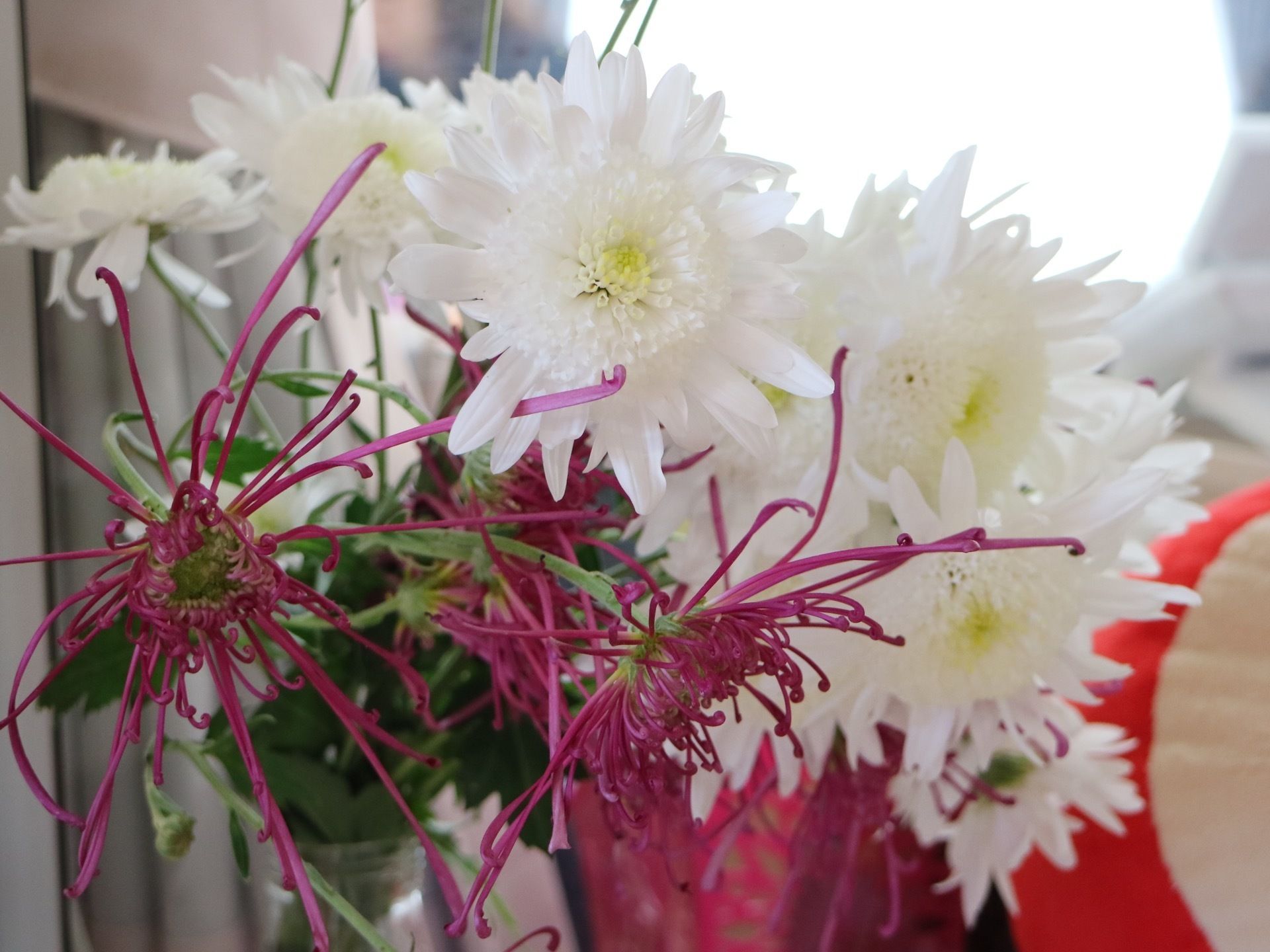 施設内の窓際に置かれたインテリアである。白色や紫色の花が約20輪、透明の花瓶に生けられて飾られている。
