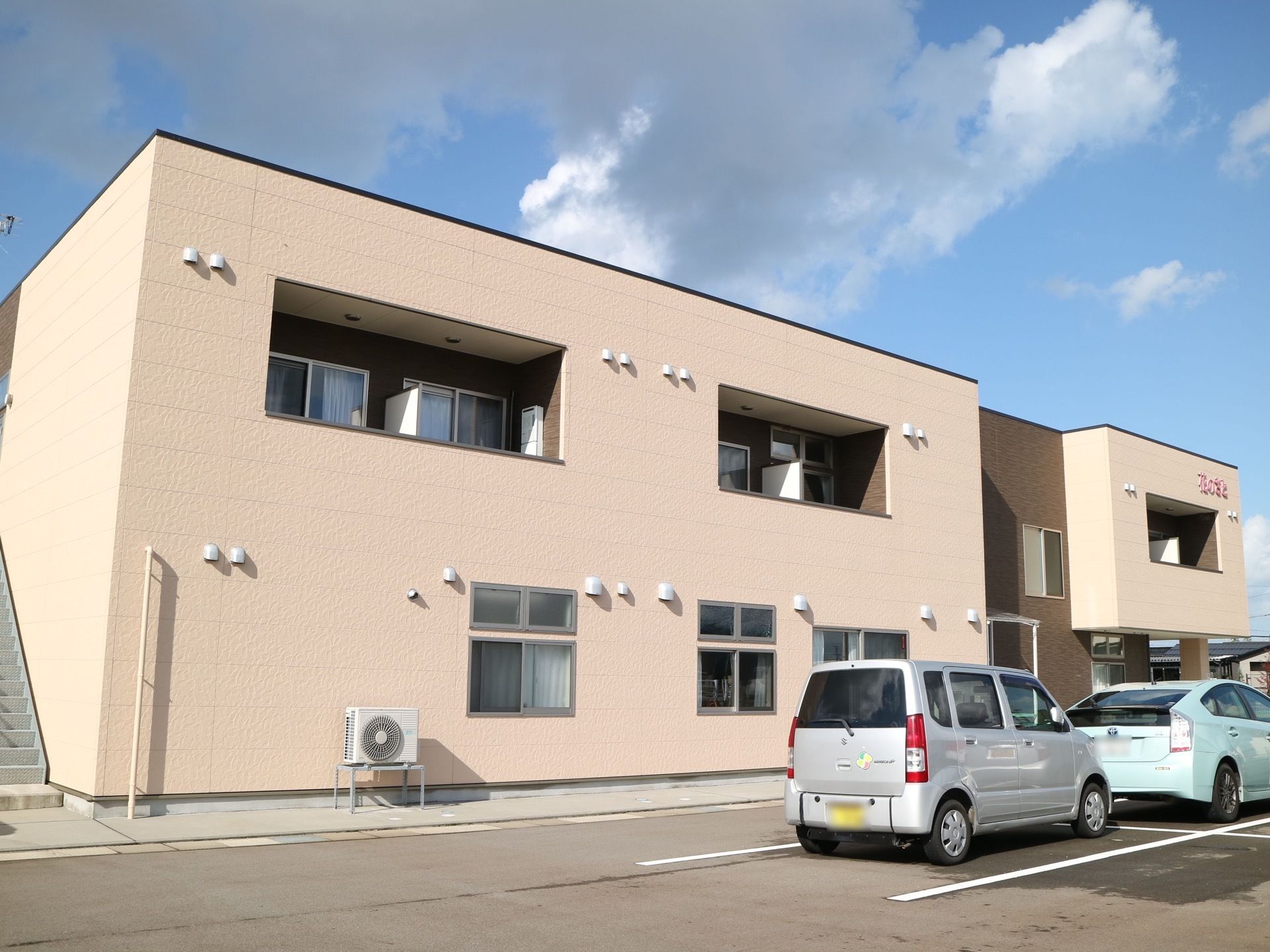 施設の建物は２階建てで、淡いベージュ色の外観である。建物の側には駐車場が設けられており、そこからは居室に備わるベランダが見える。