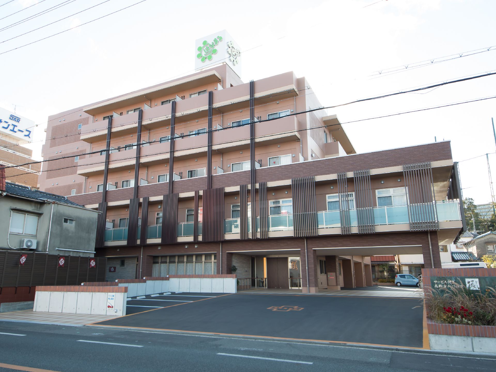 5階建ての建物があり、施設前の路面はアスファルトで舗装されている。施設名が書かれた看板があり、その手前は花壇になっている。