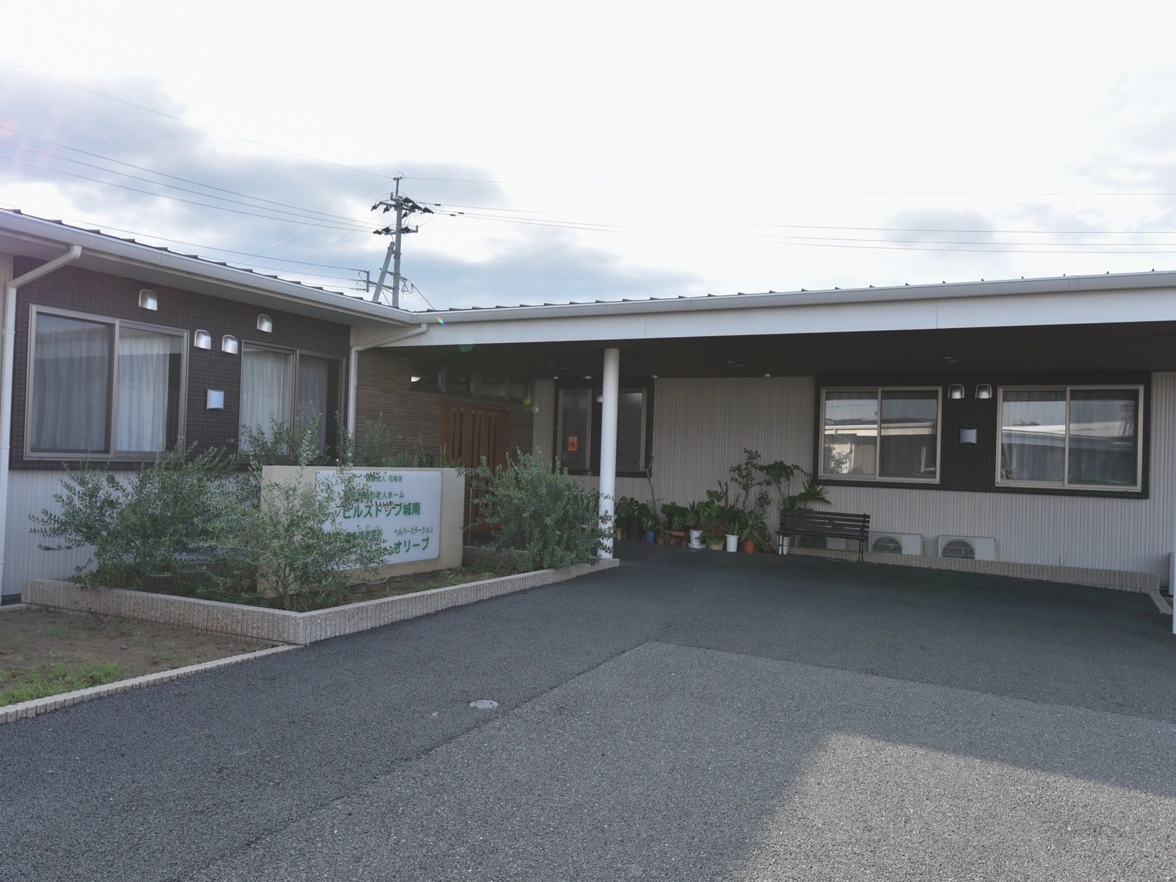 アスファルトで整備された敷地に平屋建ての建物がある。植栽スペースに施設名が表示された塀が設置されている。