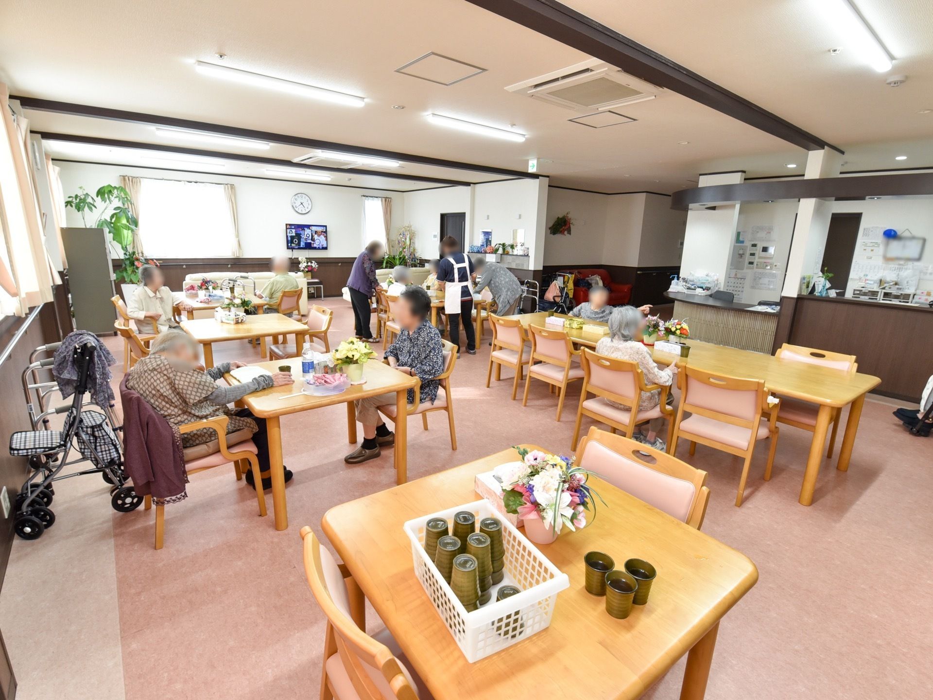 茶色いダイニングテーブルと椅子がたくさん並べて置いてある。数名の入居者様が椅子に座ったり、立ったりされている。