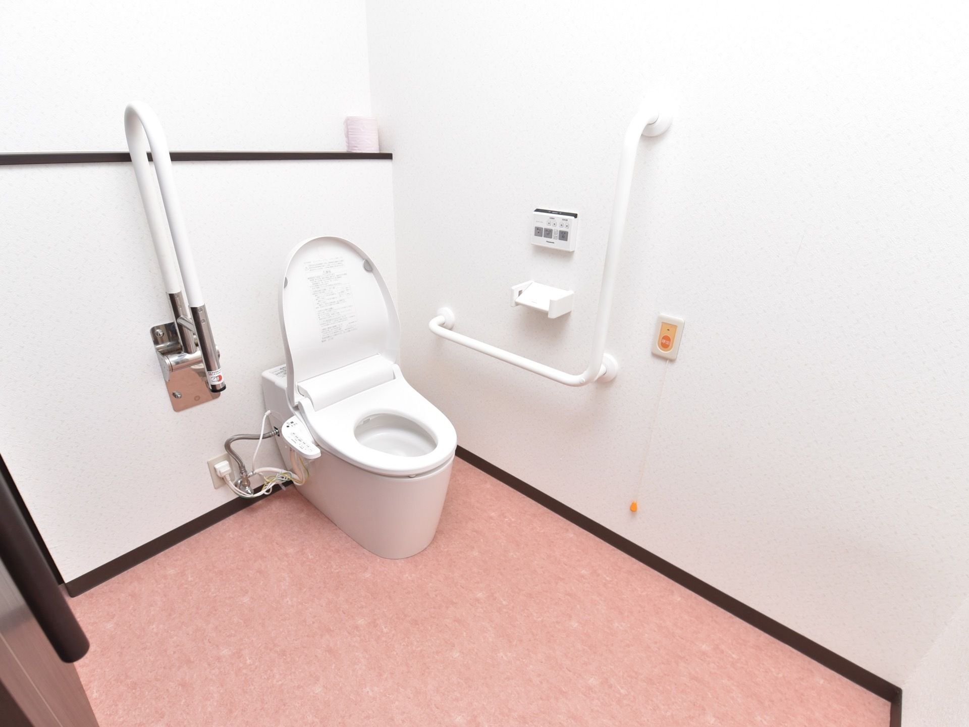 白い壁とピンク色の床の内装である。白いタンクレスのトイレの便座が開いている。壁に固定式の手すりと可動式の手すりが設置されている。