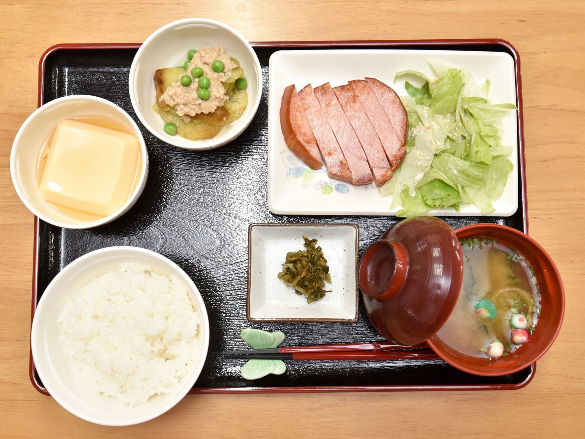 四角い黒い配膳盆におかずとみそ汁とご飯が並べて置いてある。献立は卵豆腐やハムを焼いたものなどである。