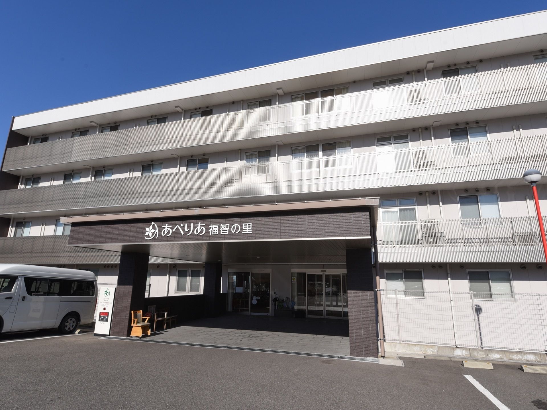 白ベースの４階建ての施設外観である。玄関前には駐車場が完備されており、入り口まで地面はフラットで段差が一切ない。