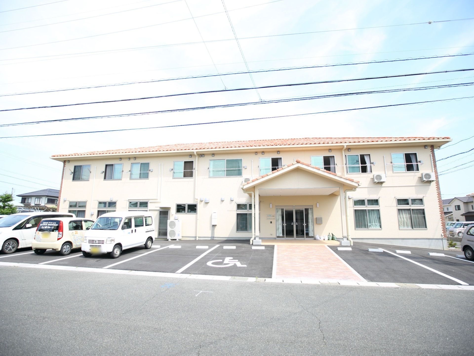 アスファルトで平らに舗装された敷地に２階建ての建物が建っている。手前には駐車場があり、車が４台ほど停められている。