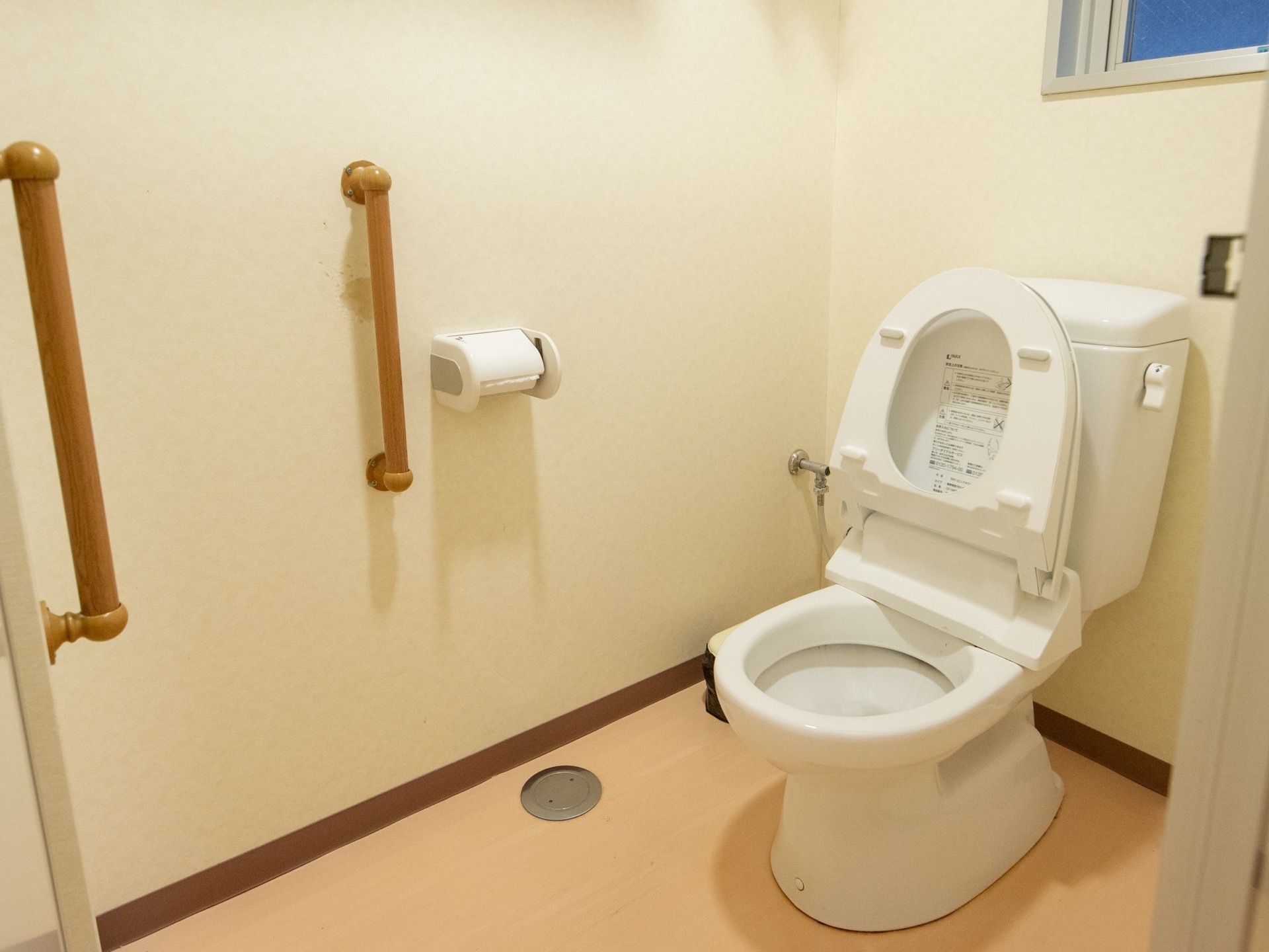 オレンジ色の床と白壁のトイレ内。洋式便器が設置されている。２か所に木目調の手すりが備えつけられている。便器の上部には窓がある。