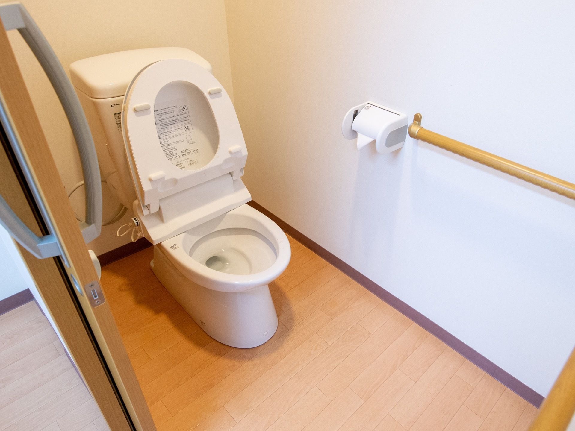 フローリングの床に、洋式トイレが設置されている。壁にはペーパーホルダーと木製の手すりがついている。扉は引き戸になっている。