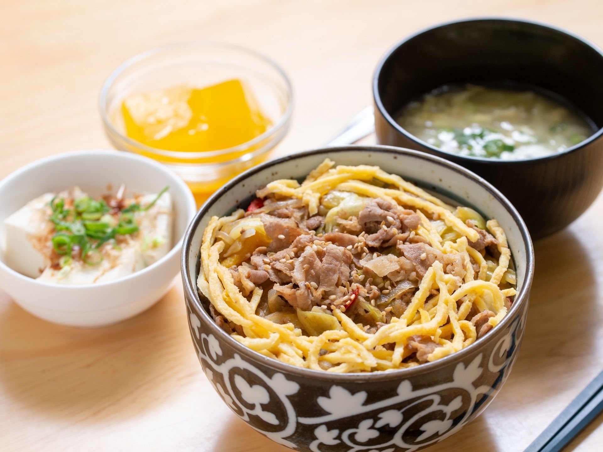 献立の一例。卵と肉のどんぶりとみそ汁、冷や奴とオレンジ色のゼリーが机に並んでいる。陶器や木製の食器を使用している。