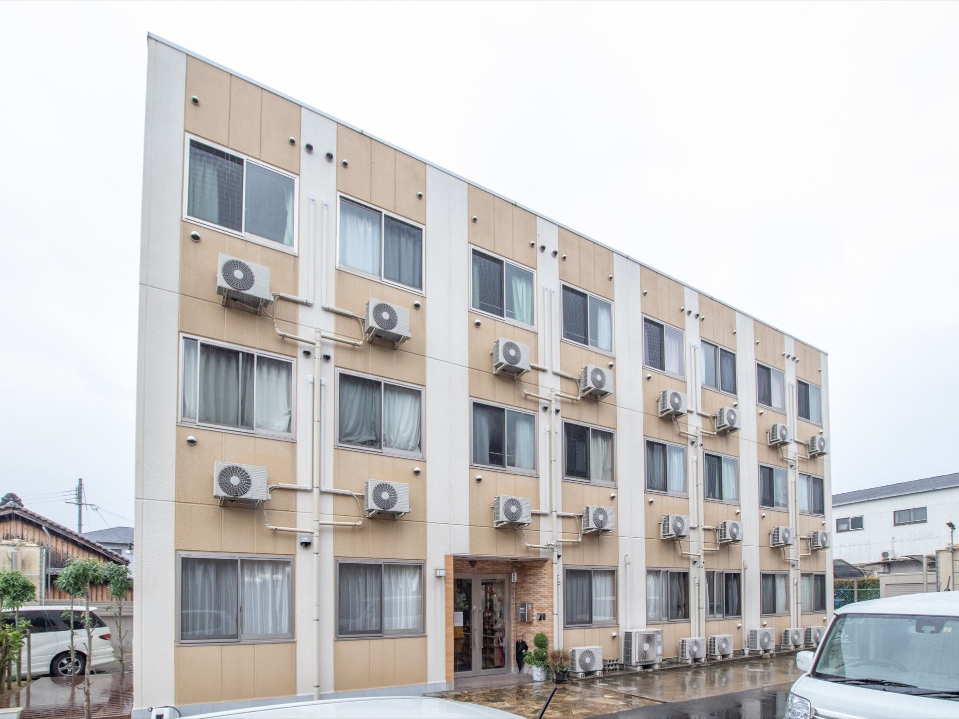 3階建ての施設建物である。中央に玄関、各部屋の壁にはエアコン室外機が設置されている。玄関前には植木鉢が置かれている。