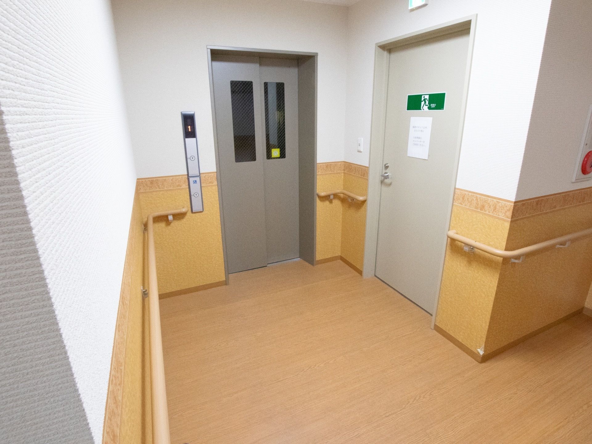 上半分が白色、下半分がライトブラウンの壁に囲われたような場所に、エレベーターと、非常階段がある。壁には、手すりが設置されている。