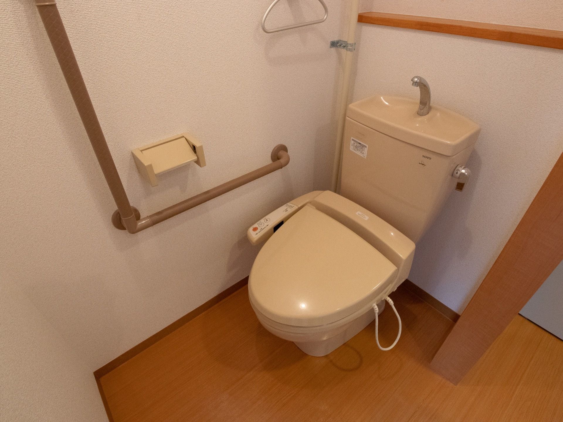 温水洗浄便座が設置されているトイレである。白い壁に木目調の床の造りである。壁には、茶色いL字型の手すりが取りつけられている。