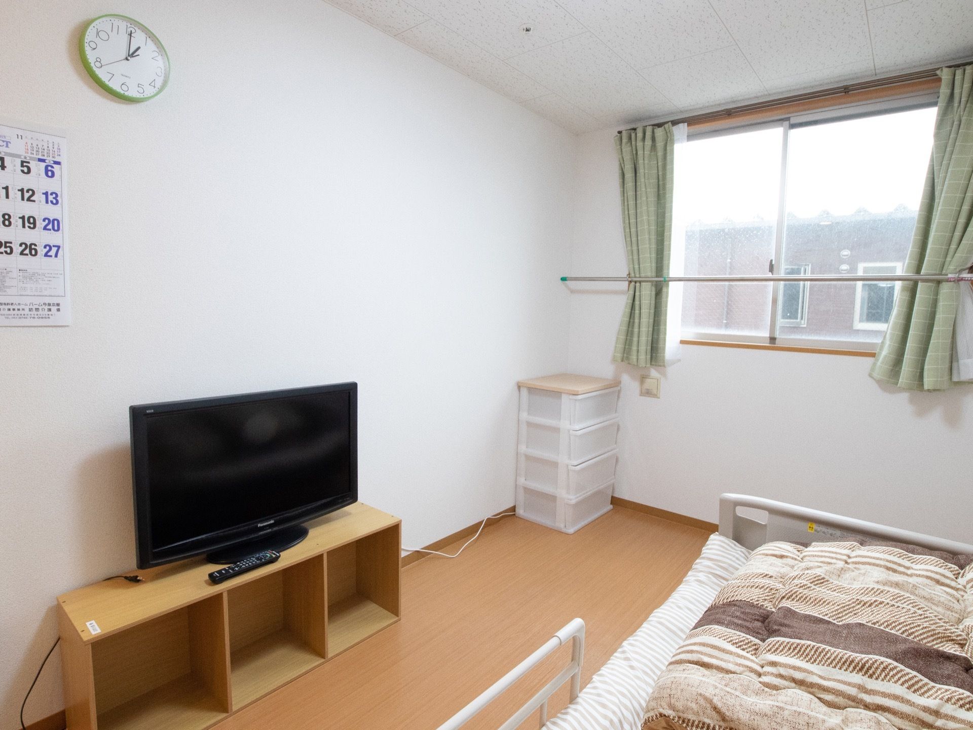 緑のカーテンが掛かっている窓から、隣の建物が見える。居室の壁は白く、床は木目調のフローリングである。テレビとベッドが置いてある。