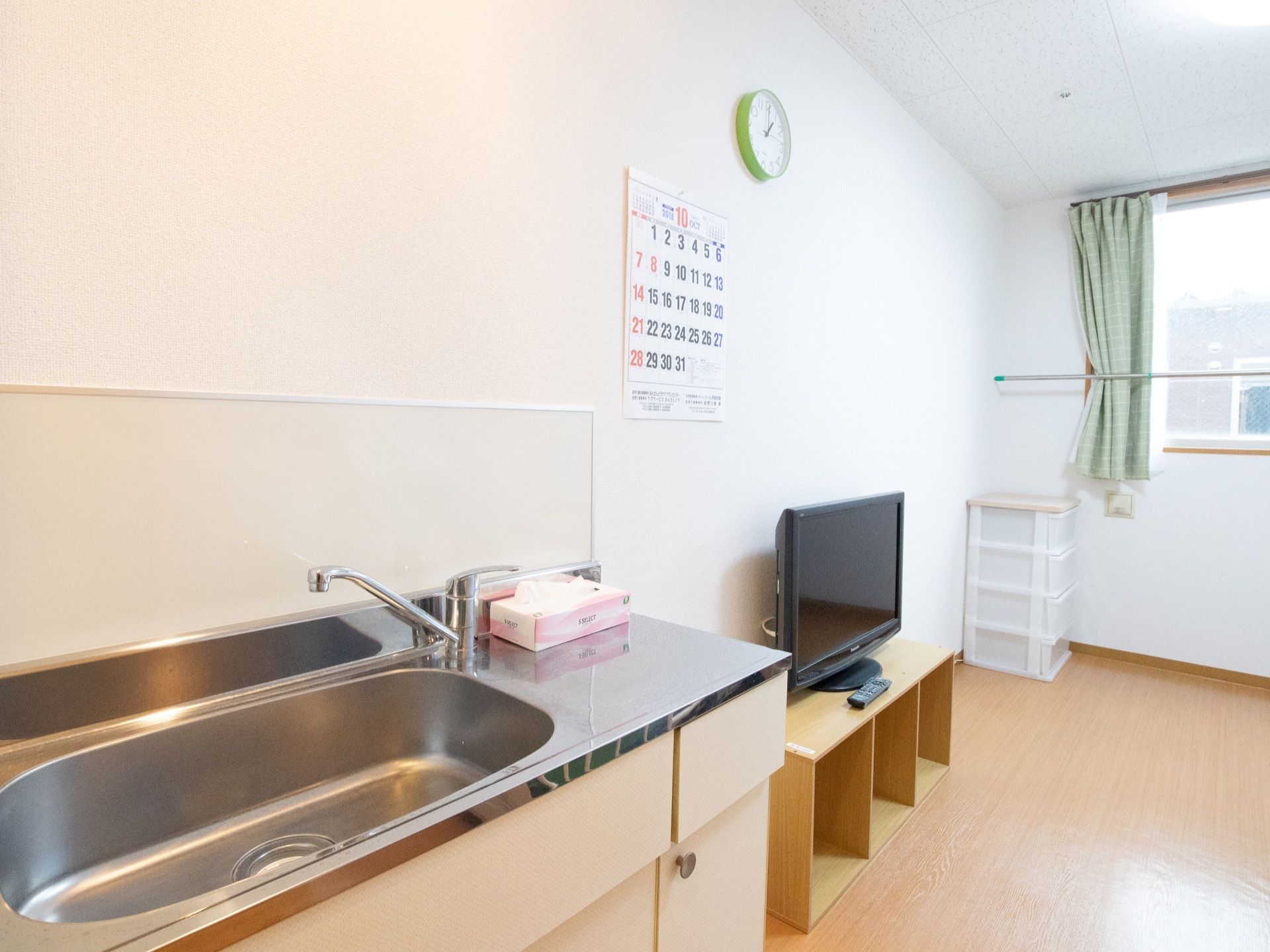 居室入り口を入ってすぐに、ミニキッチンが設置されている。壁には汚れよけのパネルが取りつけられている。テレビと収納ケースが置かれている。