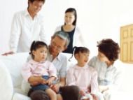白いソファに座った高齢の夫婦がピンクの服を着た孫娘2人と笑顔で会話。ソファの後ろでは子供たちの親が笑顔で見守っている。
