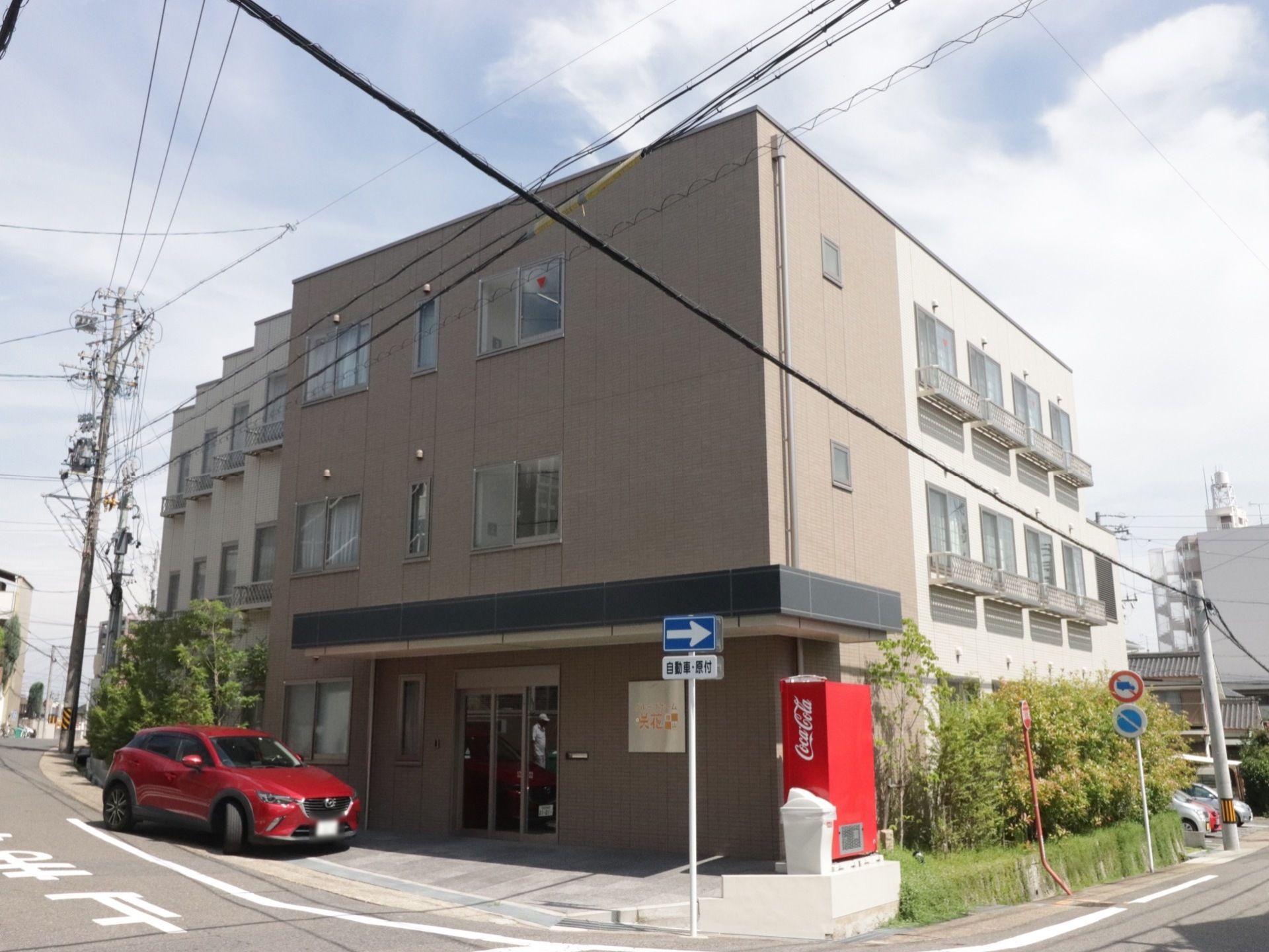 ３階建ての茶色を基調とした建物。奥には駐車スペースもあり、ご家族の方が来訪された際、使っていただける。