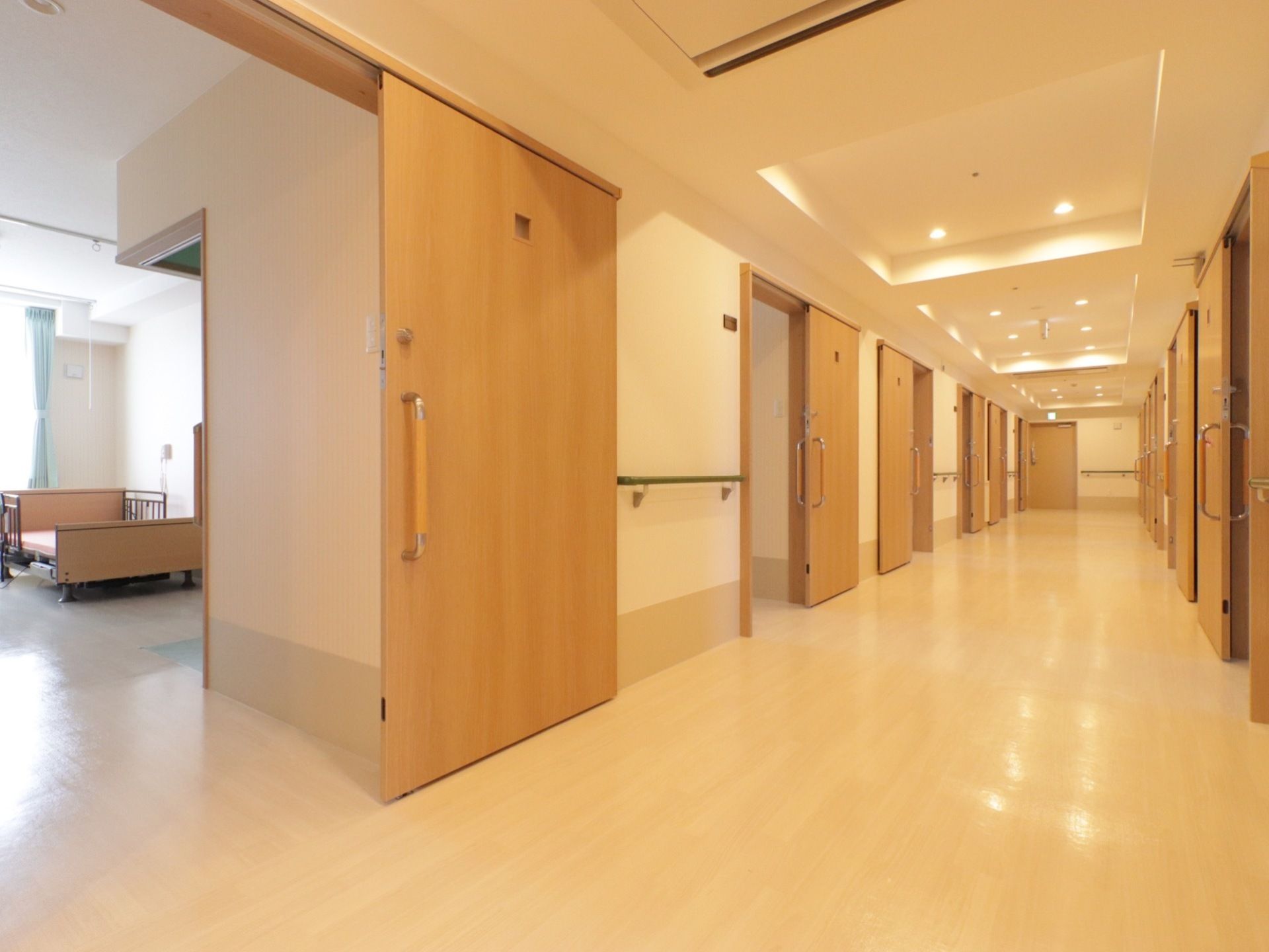 廊下に居室ドアが両側に並んでいる。ドアの間の壁には手すりが取りつけられている。ドアの横にネームプレートが貼られている。