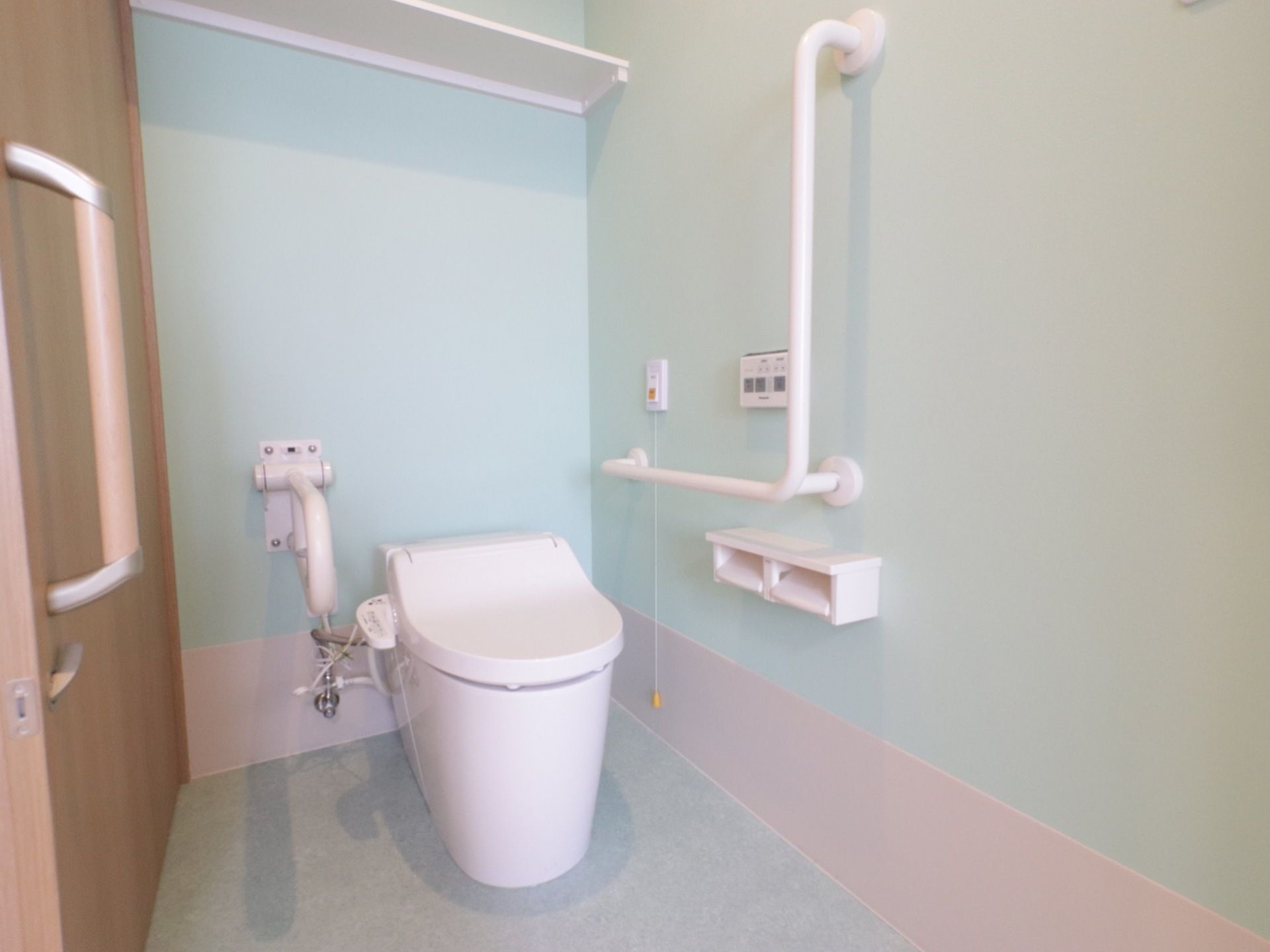 トイレの天井近くに棚が１段設置されている。壁には固定式と折り畳み式の２種類の手すりが取りつけられている。