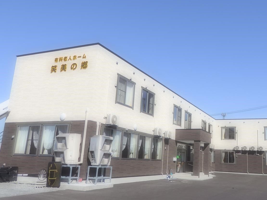 「住宅型有料老人ホーム 笑美の郷」と2階部分に書かれた2階建ての建物を左斜め前から撮影。2階部分は白、1階部分はこげ茶、玄関上には屋根がある。