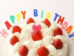 カラフルなアルファベットのロウソクで「HAPPY　BIRTHADAY」とつづられたイチゴのショートケーキ。ケーキ中央のイチゴには小さなピンクのリボンが乗っている。