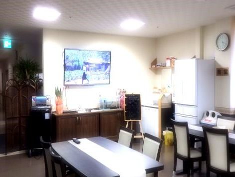中央に壁掛けテレビ、右手に冷蔵庫と壁掛けテレビ、左手にカラオケ機器、中央手前に4人掛けのテーブルが二つ並んでいる。