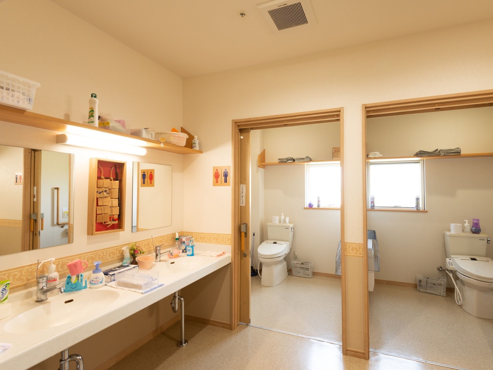 トイレの個室が2つ並んで設置されている。左手前に洗面台と鏡が取りつけられており、照明の上方に棚がある。