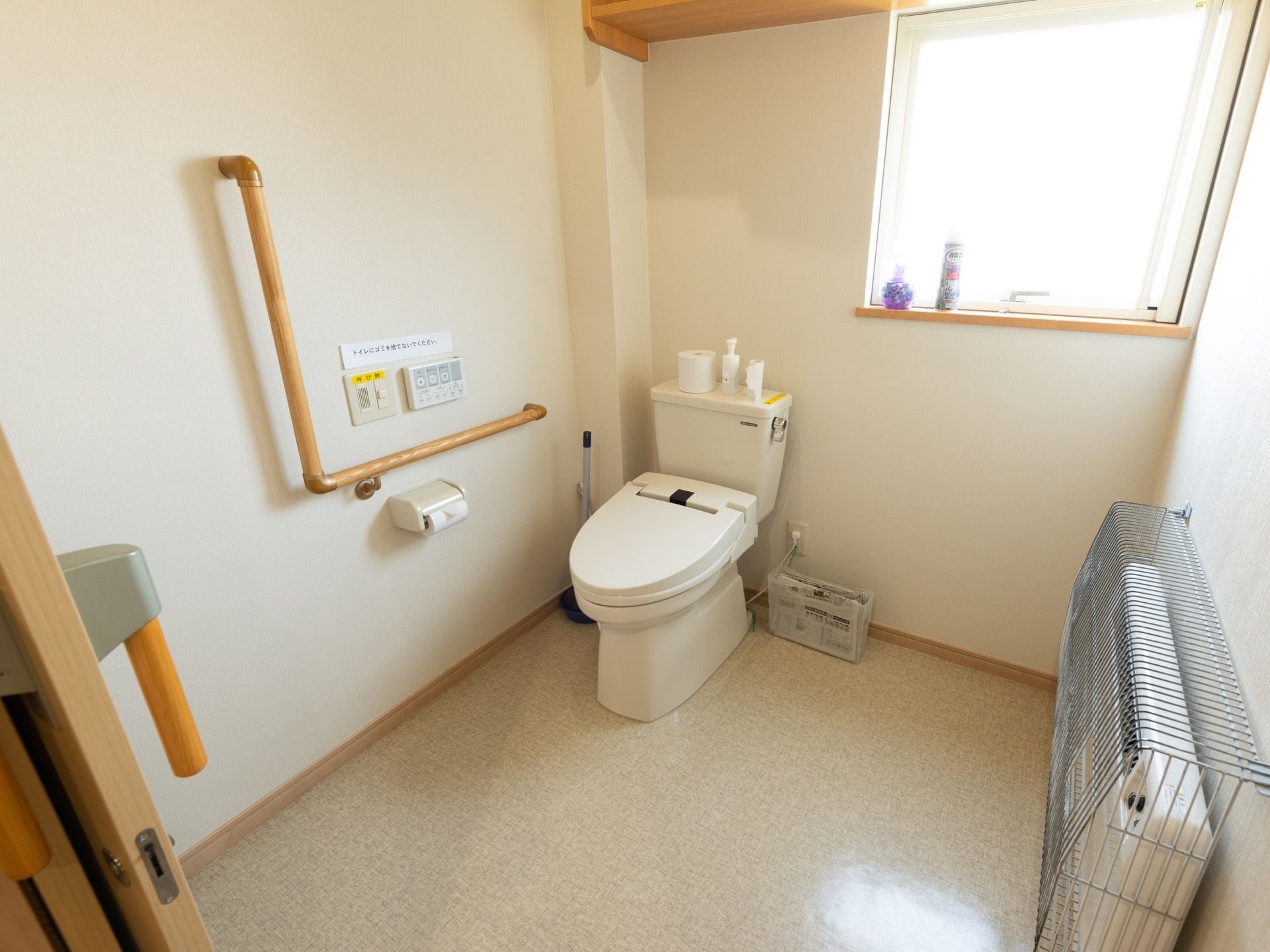 大きな窓がついたトイレスペースに、洋式便器がある。右側の壁面に暖房器具が設置されており、安全カバーが施されている。