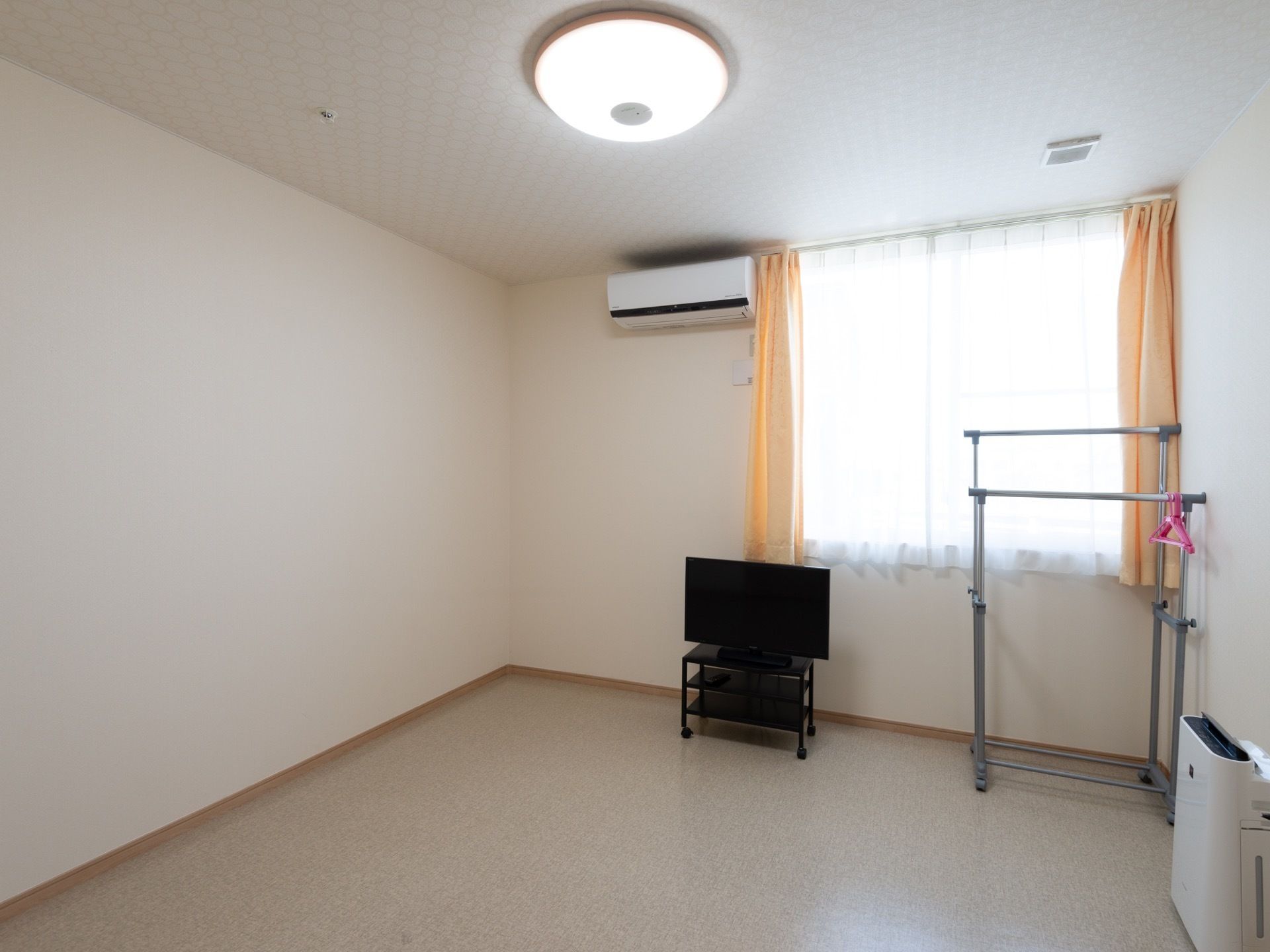 ハンガーラックやテレビ台、空気清浄機が置かれている個室である。窓側の壁面にエアコンが設置されている。